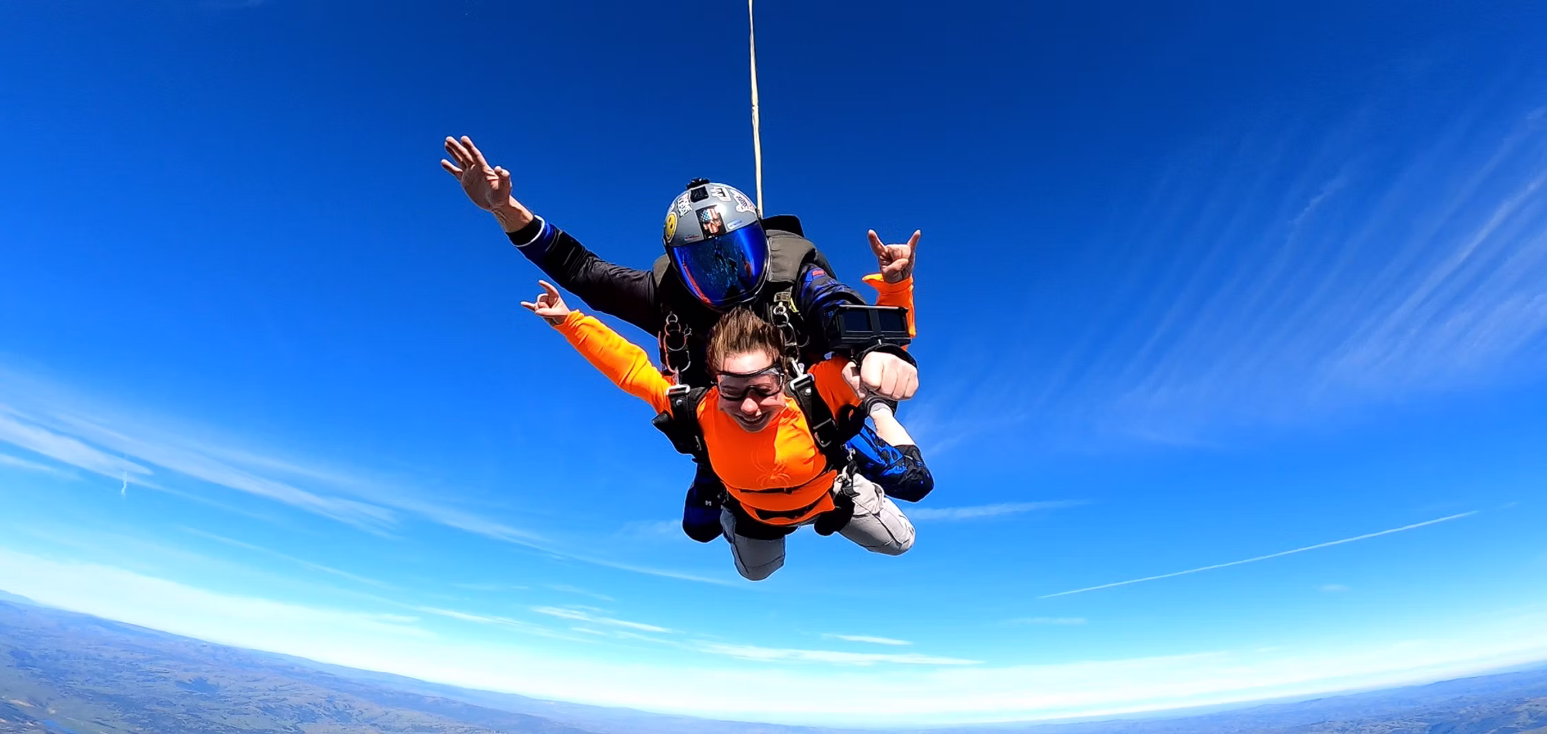 Tandem jumper throwing a shaka in freefall under a deep blue sky.